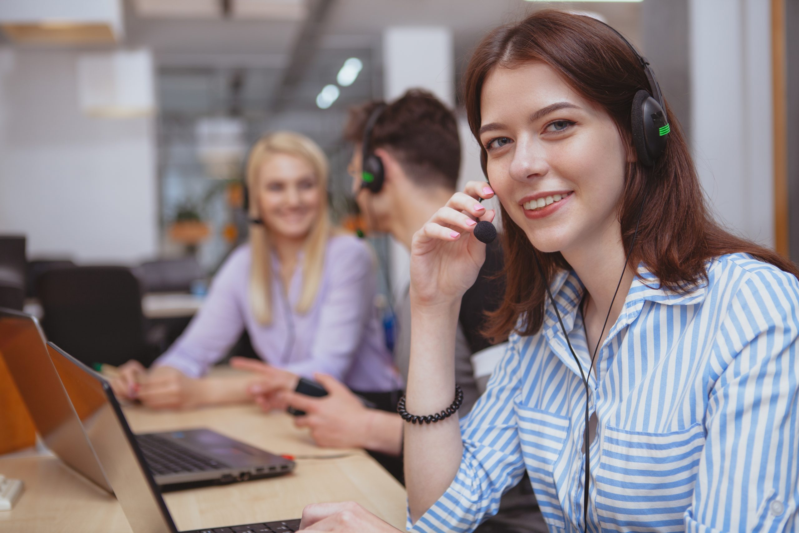 Cheerful helpful female call center operator smiling to the camera, working at the office, copy space. Charming young woman working at customer support service. Happy female at technical support office answering clients calls