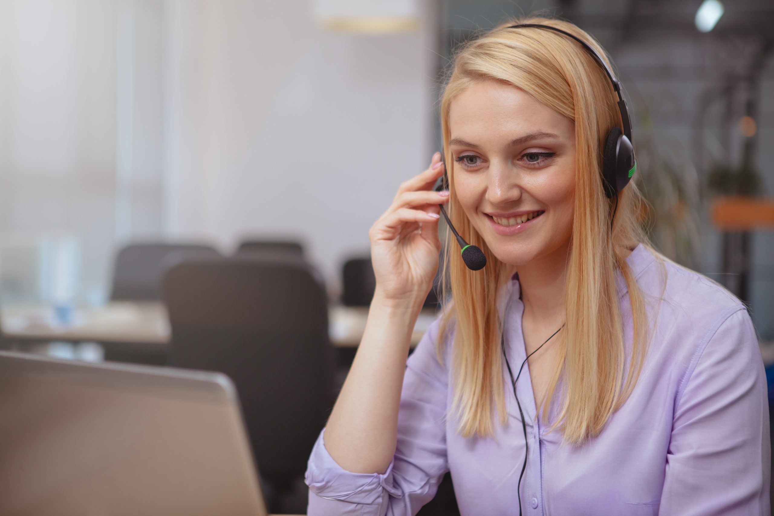 Beautiful cheerful woman smiling joyfully, answering customer calls at the call center, typing on her laptop. Lovely female customer support operator talking with callers, wearing headset