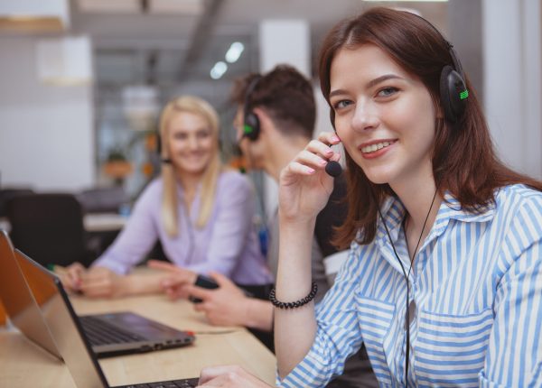 Cheerful helpful female call center operator smiling to the camera, working at the office, copy space. Charming young woman working at customer support service. Happy female at technical support office answering clients calls